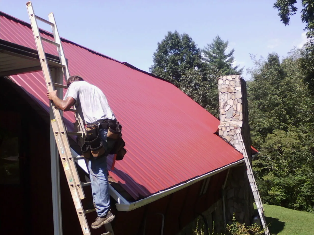 Roofer on a ladder beside a red metal roof and stone chimney — Soffit & Fascia Repair by Roofing Brothers in Green Valley Lake